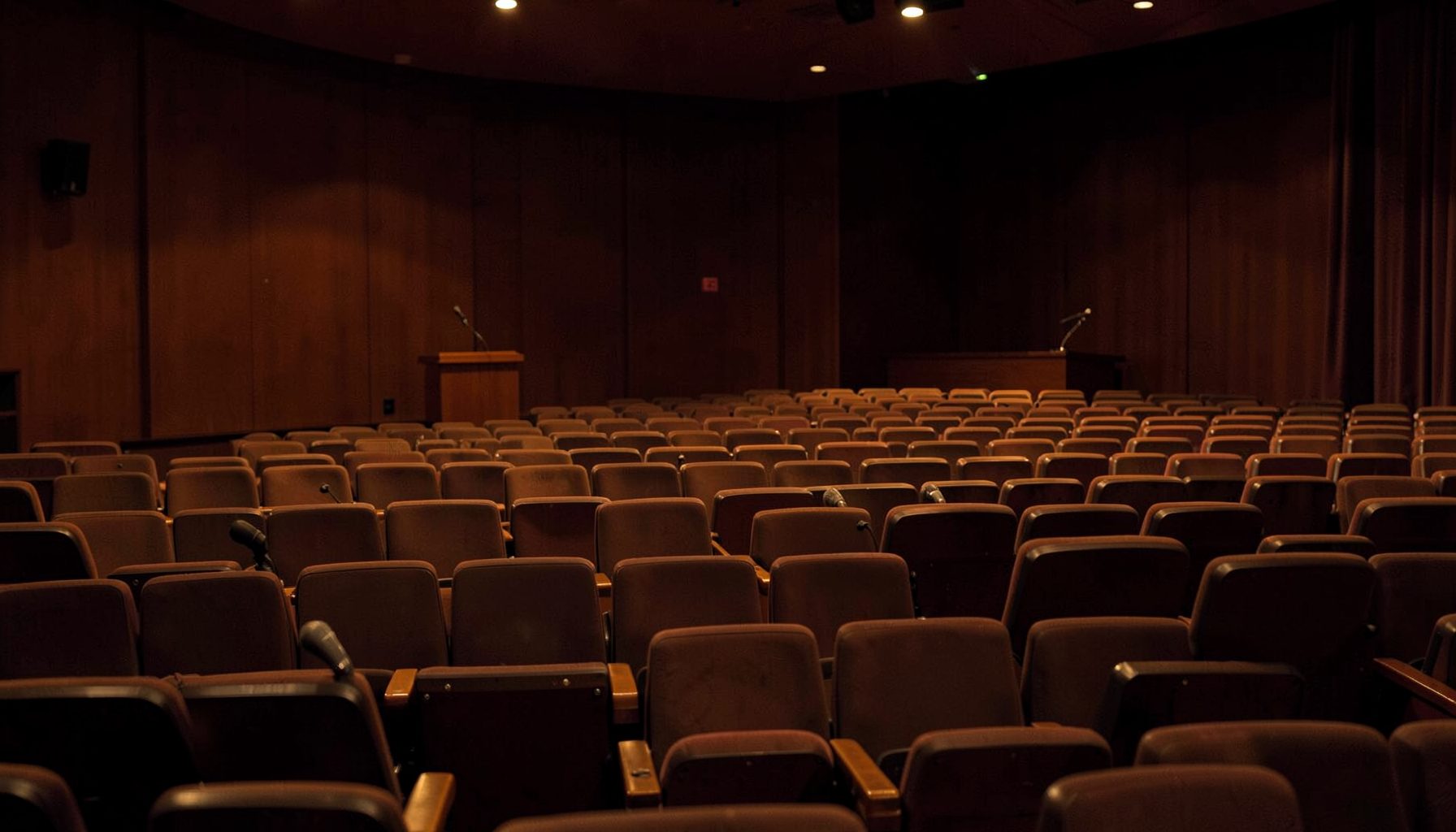 A warmly lit auditorium with rows of empty seats and a lectern on stage, deep amber and brown tones, quiet and dignified atmosphere